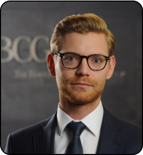 A person with reddish-brown hair and a beard, wearing black-rimmed glasses, a dark suit, and a tie, with a confident expression, standing in front of a backdrop with the letters "BCG".