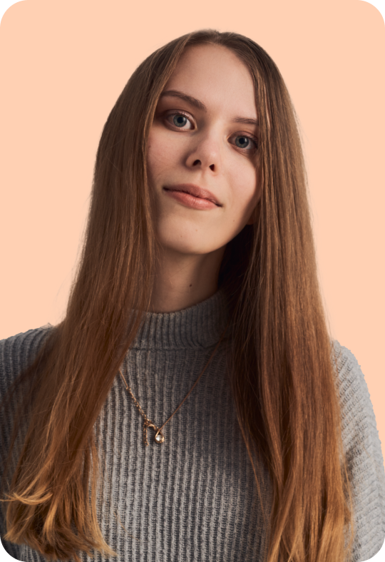 A person with long straight brown hair, wearing a grey sweater and a delicate necklace, looking straight at the camera with a soft smile, against a pale orange background.