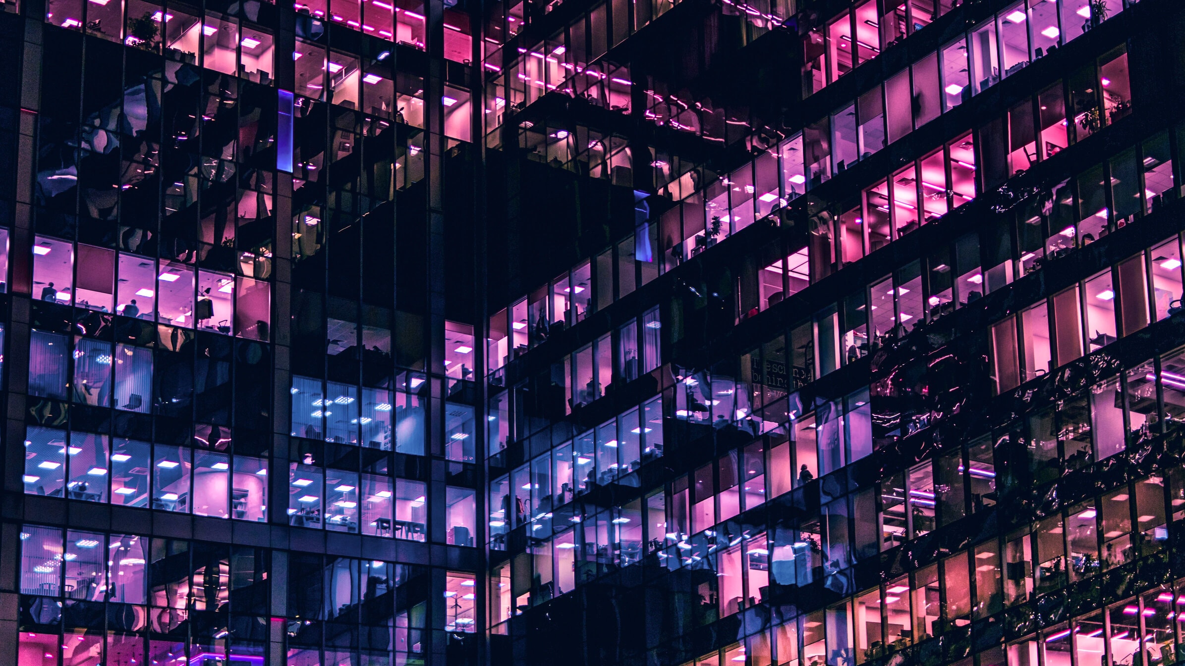 An artistic photo of a building during the night. The modern building is mostly made of glass that reflects purple, pink and blue colours of the night city.